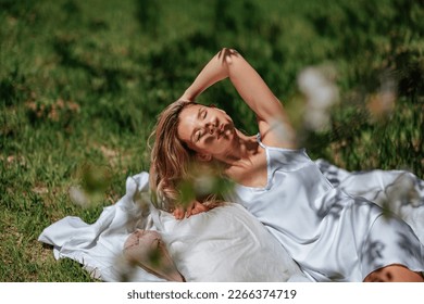 woman sleeps on a white bed in the fresh spring grass in the garden. Dressed in a blue nightgown.