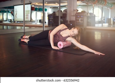 Woman In Sleeveless Shirt And Black Leggings In Gym Doing Exercise With Foam Or Fascia Roll And Looking At Camera