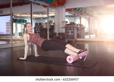 Woman In Sleeveless Shirt And Black Leggings In Gym Doing Exercise With Foam Or Fascia Roll And Looking At Camera