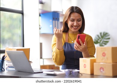 Woman in a small start-up business woman working in a home office working on a laptop and smartphone checking orders from the internet in preparation for shipping. - Shutterstock ID 2202619041