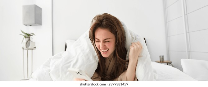 Woman with smartphone, feeling excitement, reading mobile phone and cheering, lying in bed under white blanket, using smartphone and looking happy in morning. - Powered by Shutterstock