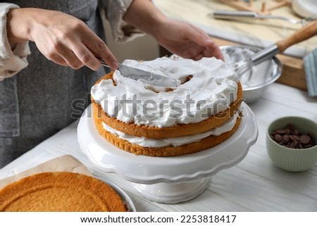 Woman smearing sponge cake with cream at white wooden table in kitchen, closeup