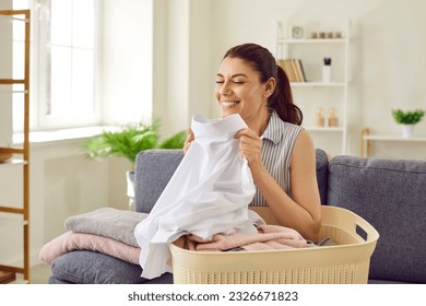 Woman smelling clean laundry. Happy beautiful young housewife sitting on couch with laundry basket, holding perfectly clean, washed, white shirt, smelling fresh, natural aroma, and smiling - Shutterstock ID 2326671823