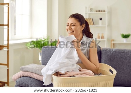 Woman smelling clean laundry. Happy beautiful young housewife sitting on couch with laundry basket, holding perfectly clean, washed, white shirt, smelling fresh, natural aroma, and smiling