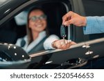 A woman with a smile and an outstretched hand receives car keys from a man in a blue suit, depicting a moment of successful customer interaction within a well-lit car dealership.