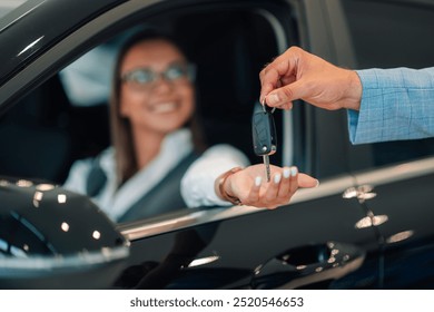 A woman with a smile and an outstretched hand receives car keys from a man in a blue suit, depicting a moment of successful customer interaction within a well-lit car dealership.