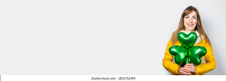 Woman with a smile in a yellow jacket and a scarf holds a clover balloon on a white background. Concept of a holiday, celebration, party, St. Patrick's Day, Ireland. Banner - Powered by Shutterstock