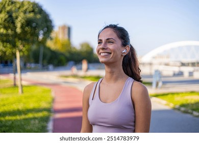 A woman smiles brightly while enjoying an outdoor workout. She wears a tank top and earbuds, representing health, fitness, and happiness in a sunny park setting. - Powered by Shutterstock