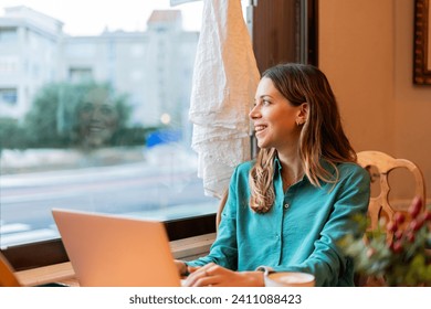 A woman smiles as she works on her laptop at a vintage café table by the window, embodying the remote work lifestyle
