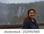 A woman smiles while standing in front of a misty waterfall at Plitvice Lakes National Park, Croatia, on a cloudy day. The scenic landscape adds depth to the portrait.