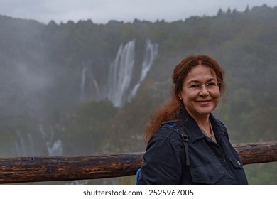 A woman smiles while standing in front of a misty waterfall at Plitvice Lakes National Park, Croatia, on a cloudy day. The scenic landscape adds depth to the portrait. - Powered by Shutterstock