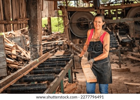 Woman smiles and winks one eye looking at camera, loading a board in a sawmill. Despite the hardships, women in these roles continue to demonstrate their resilience and determination to get ahead.