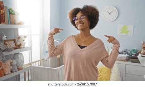 Woman smiling with curly hair points to herself in a cozy bedroom with a baby cradle surrounded by soft toys and pastel decor, radiating warmth and style.