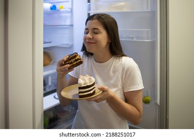 Woman Smiling Enjoys A Delicious Sweet Chocolate Cake At Night Alone Near The Fridge. Funny Photo. A Woman With Trouble Eating. Carbohydrates, Calories. Nocturnal Harmful Forbidden Food