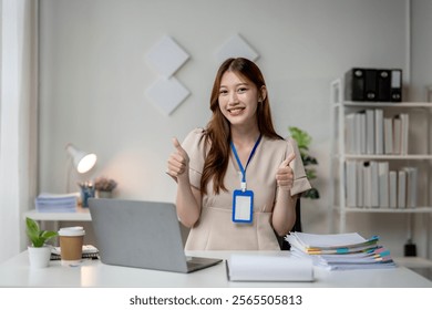 A woman is smiling and giving a thumbs up while sitting at a desk with a laptop and papers
