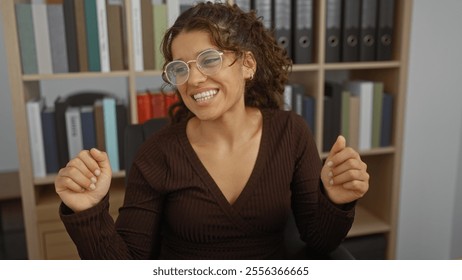 Woman smiling indoors, brunette, wearing glasses, sitting in office, surrounded by bookshelves, work environment, happy expression, casual attire, adult female, attractive hispanic - Powered by Shutterstock