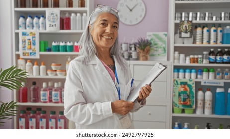 Woman smiling in a pharmacy, grey-haired, wearing a white lab coat while holding a clipboard, surrounded by shelves stocked with various pharmaceutical products - Powered by Shutterstock