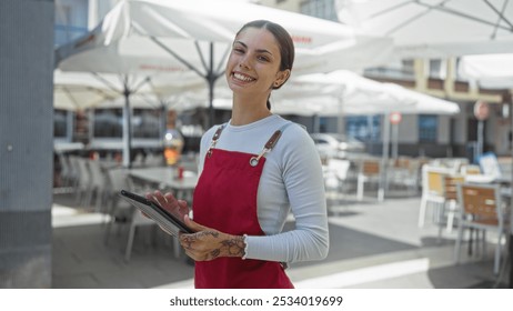 Woman smiling in red apron standing on outdoor cafe terrace and holding tablet on sunny day with white umbrellas in background - Powered by Shutterstock