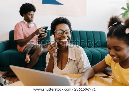 Woman smiling and speaking on the phone while sitting next to her daughter, who is using a laptop. Happy, modern family using different technology devices in their leisure time at home.