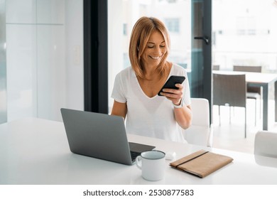 Woman Smiling While Engaging with Phone and Laptop at Kitchen Island - Powered by Shutterstock