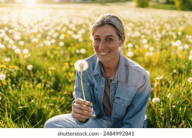 woman smiling while holding a dandelion in a sunny field full of dandelions, showcasing the beauty of nature in spring. The sun is shining brightly in the background. - Powered by Shutterstock