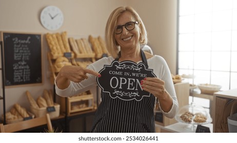 Woman smiling while pointing at open sign in cozy bakery with fresh baked goods and rustic decor - Powered by Shutterstock