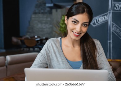 Woman smiling while typing on silver laptop at wooden café table with chalkboard art and plant. Co-working, café, workspace, modern, relaxed, professional, creativity