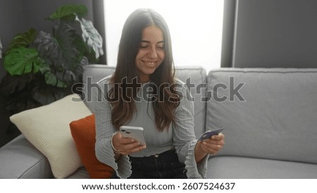 Woman smiling while using smartphone and credit card on couch in cozy living room, showcasing modern technology and relaxed home lifestyle.