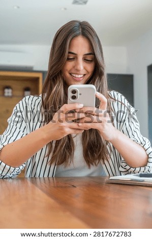 Woman smiling while using smartphone at home, texting and browsing social media at wooden table