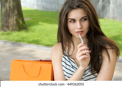 Woman smokes electronic cigarette. She sits in a park with shopping bags.