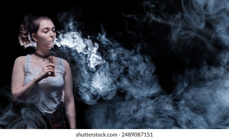 Woman smokes vape. Girl is engaged in vaping. Beautiful lady vaper. Female student exhales smoke from vape. Electronic cigarette in hand of vaper. Woman smokes steam cigarette. Vaper stands in dark