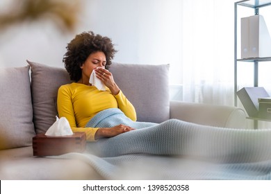 Woman sneezing in a tissue in the living room.  Woman blowing her nose on couch at home in the living room. African American woman using a tissue sitting on a sofa at home
