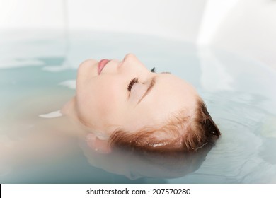 Woman soaking in a bath tub with just her face above the water with her eyes closed and a beautiful serene expression