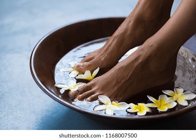 Woman soaking feet in bowl of water with floating frangipani flowers at spa. Closeup of a female feet at wellness center on pedicure procedure. Woman feet in spa wooden bowl with exotic white flowers.