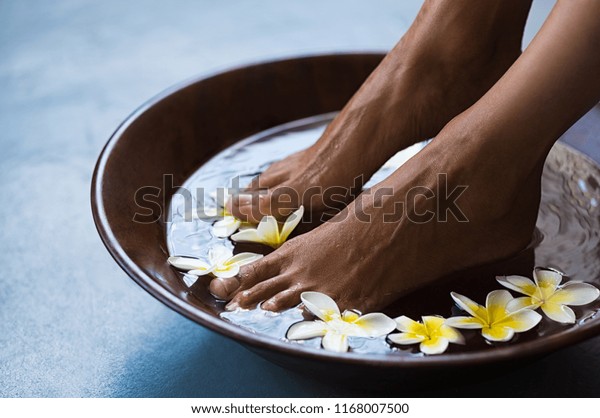 Woman soaking feet in bowl of water with\
floating frangipani flowers at spa. Closeup of a female feet at\
wellness center on pedicure procedure. Woman feet in spa wooden\
bowl with exotic white\
flowers.