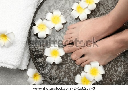Woman soaking her feet in bowl with water and flowers on light grey floor, top view. Spa treatment