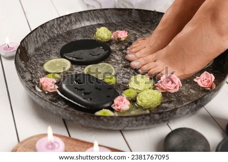 Woman soaking her feet in plate with water, stones, flowers and lime slices on white wooden floor, closeup. Pedicure procedure