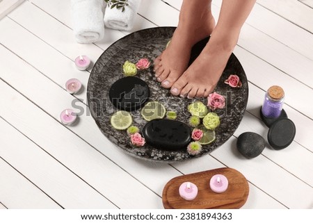 Woman soaking her feet in plate with water, stones, flowers and lime slices on white wooden floor, above view. Pedicure procedure