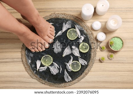 Woman soaking her feet in plate with water, flower petals and lime slices on wooden floor, top view. Pedicure procedure