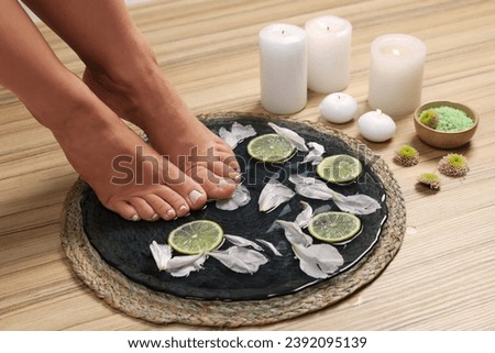Woman soaking her feet in plate with water, flower petals and lime slices on wooden floor, closeup. Pedicure procedure