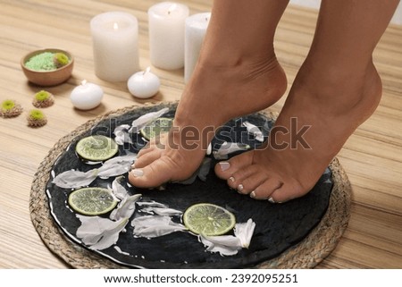 Woman soaking her feet in plate with water, flower petals and lime slices on wooden floor, closeup. Pedicure procedure