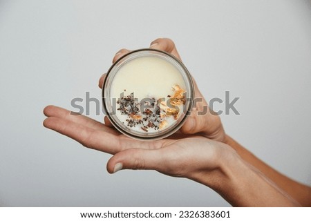 Woman soft hands holding up organic wax candle with dried plants and ingredients in round glass jar packaging. Isolated on grey background