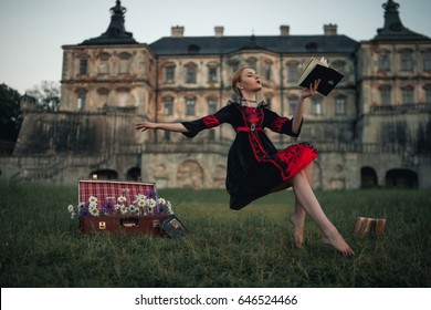 Woman sorceress flies in air and reads book against backdrop of ancient castle. Levitation. On grass there is suitcase with flowers.