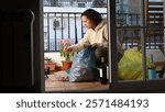 Woman sorting recycling into a blue plastic bag on her balcony. Empty wine bottles are visible near the bag.
