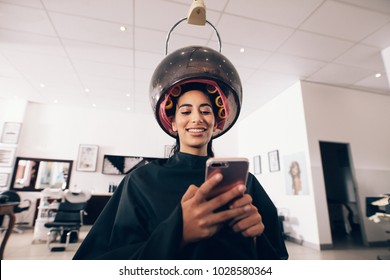 Woman At The Spa With Rollers To Her Hair And A Steaming Machine Around The Head. Smiling Woman Looking At The Mobile Phone At The Beauty Parlor While Undergoing Hair Steaming.