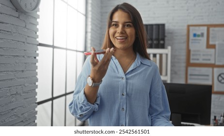 Woman speaking in an office setting with a smile, appearing professional and confident with long brunette hair and a blue blouse against a modern indoor backdrop of white brick walls and large windows - Powered by Shutterstock