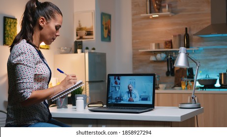 Woman Speaking Online With Doctor Taking Notes During Tele Health Sitting In Home Kitchen. Sick Lady Discussing During Virtual Consultation About Symptoms Holding Notebook And Writing Treatment