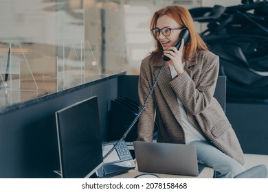 Woman In Spectacles Working In Office Wearing Casual Clothes, Talking On Phone While Sitting On Her Desk, Modern And Comfortable Office Environment, Blurred Background. Happy Employee At Work Concept