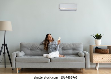 Woman spend time at home seated on sofa holding remote control manages degrees uses air conditioner enjoy air-conditioned contemporary flat, reduces heat or cold weather to comfortable temperature - Powered by Shutterstock