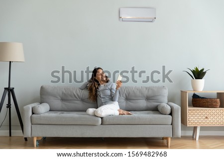 Woman spend time at home seated on sofa holding remote control manages degrees uses air conditioner enjoy air-conditioned contemporary flat, reduces heat or cold weather to comfortable temperature